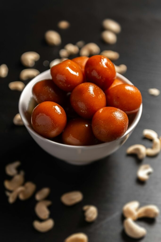 Close-up of fresh gulab jamun in a white bowl surrounded by scattered cashew nuts on a dark background.  gulab jamun recipe