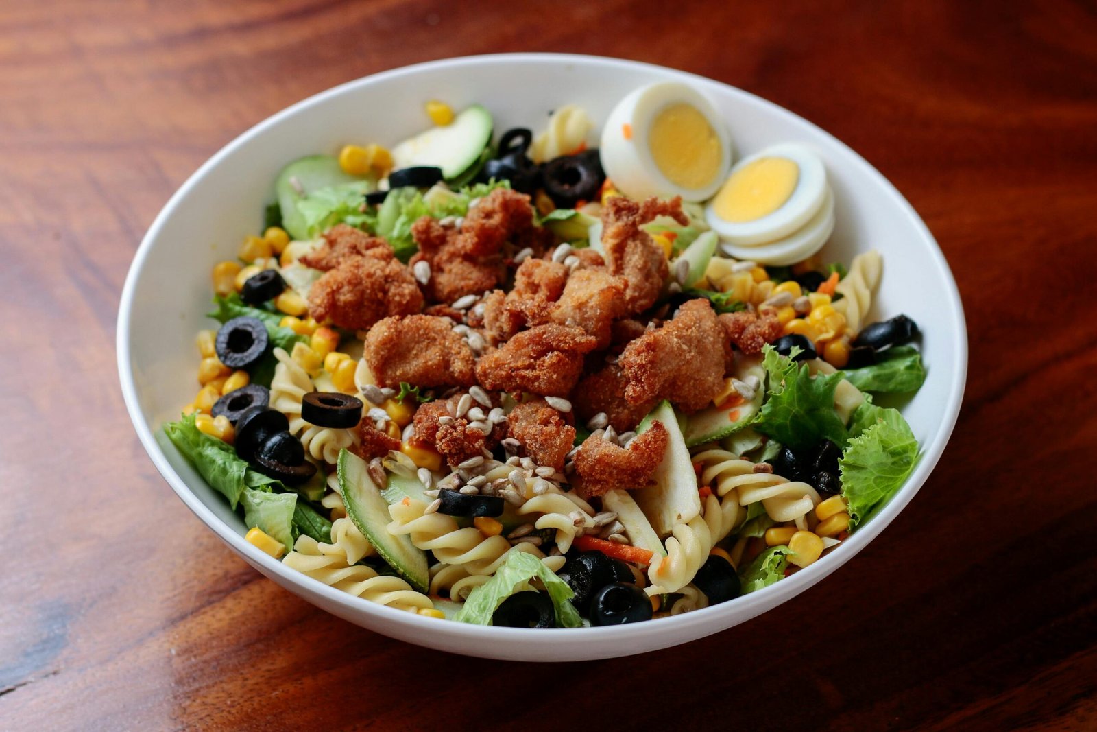 A vibrant bowl of pasta salad topped with crispy chicken, fresh vegetables, and boiled eggs.