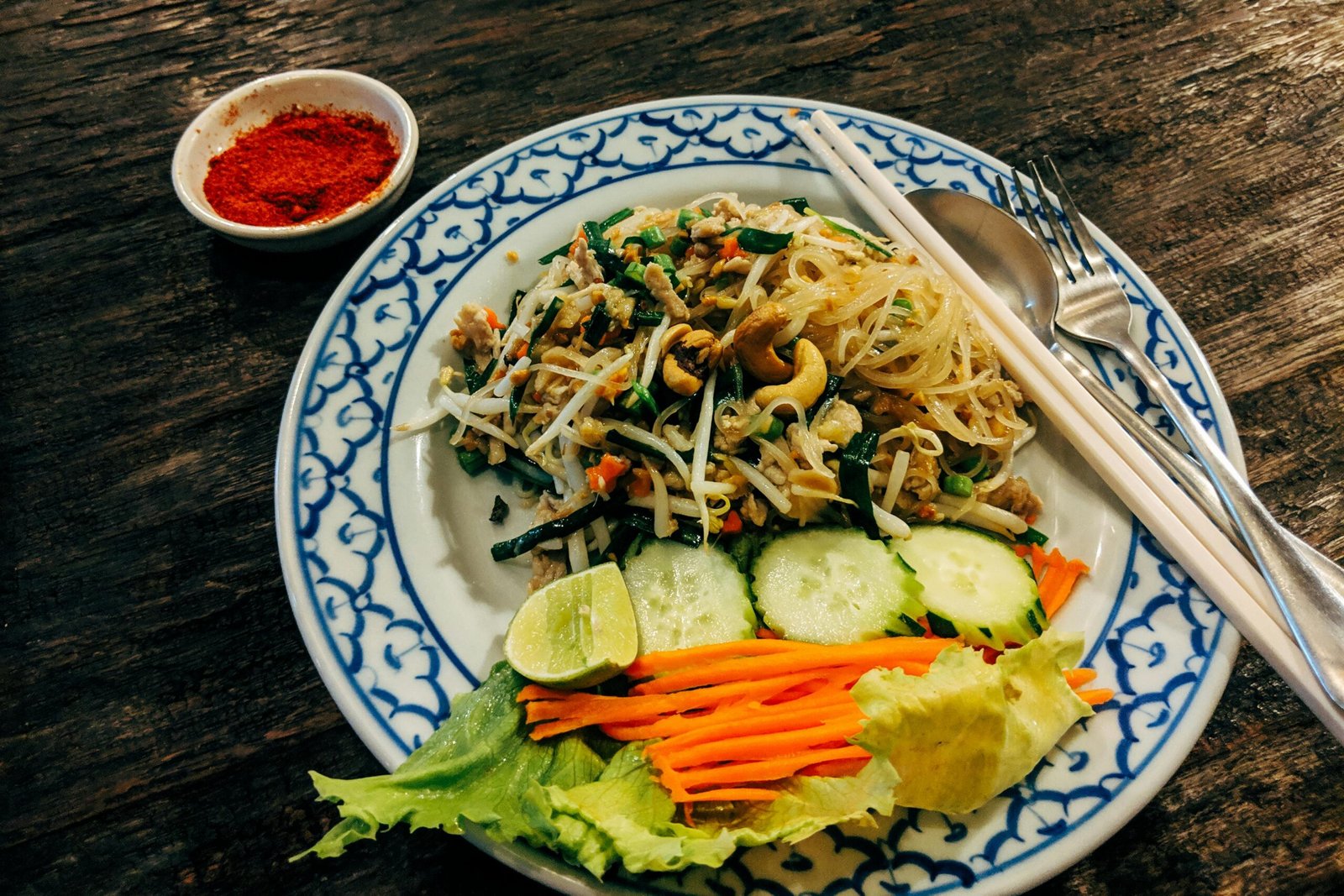 A beautifully plated Asian noodle salad with fresh vegetables and a side of chili powder.