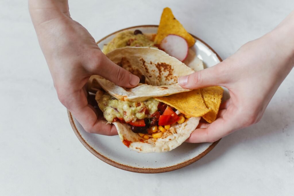 Close-up of hands holding a Mexican taco with nachos, guacamole, and colorful veggies.