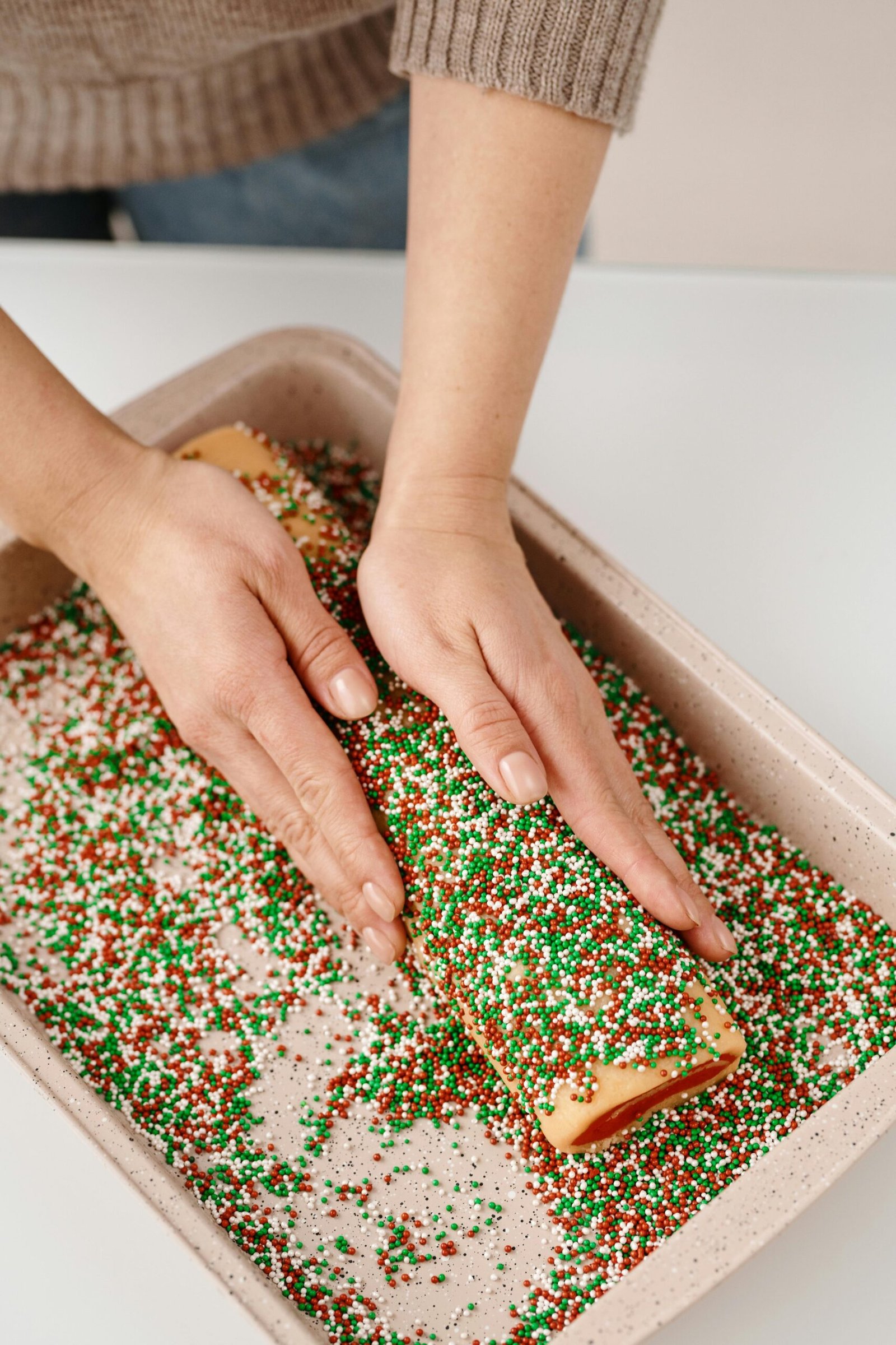 A person decorating a Christmas roll with colorful sprinkles in the kitchen.