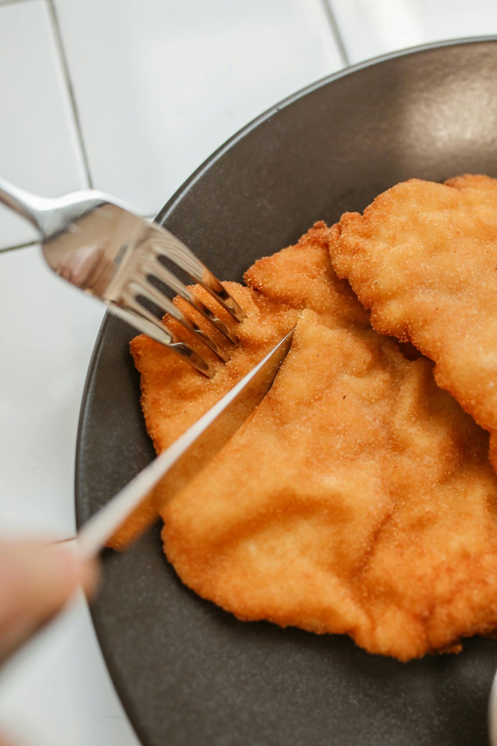 Close-up of fried schnitzel being sliced with utensils on a ceramic plate.